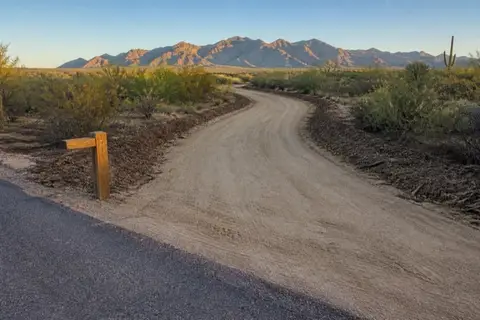 Fresh graded dirt driveway curving through cleared creosote toward desert acreage, mulched shoulders on both sides and the White Tank Mountains beyond