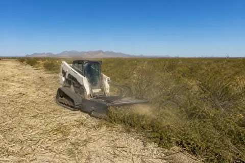 Skid-steer brush cutter working across a creosote flat, cut stubble on one side and waist-high brush on the other with the White Tank Mountains on the horizon