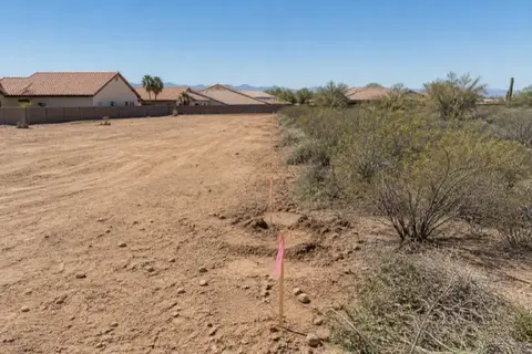 Half-cleared quarter-acre subdivision lot with smooth pale dirt on one side, creosote and young mesquite on the other, and surveyor stakes with pink flagging